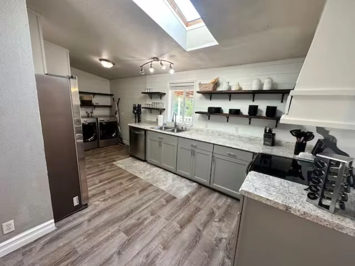 A bright and contemporary kitchen space featuring striking granite countertops, open shelving with dishware and accessories, a range of modern kitchen appliances, and a skylight that bathes the room in natural sunlight. The spacious counters and open layout suggest plenty of room for group cooking and meal prep.