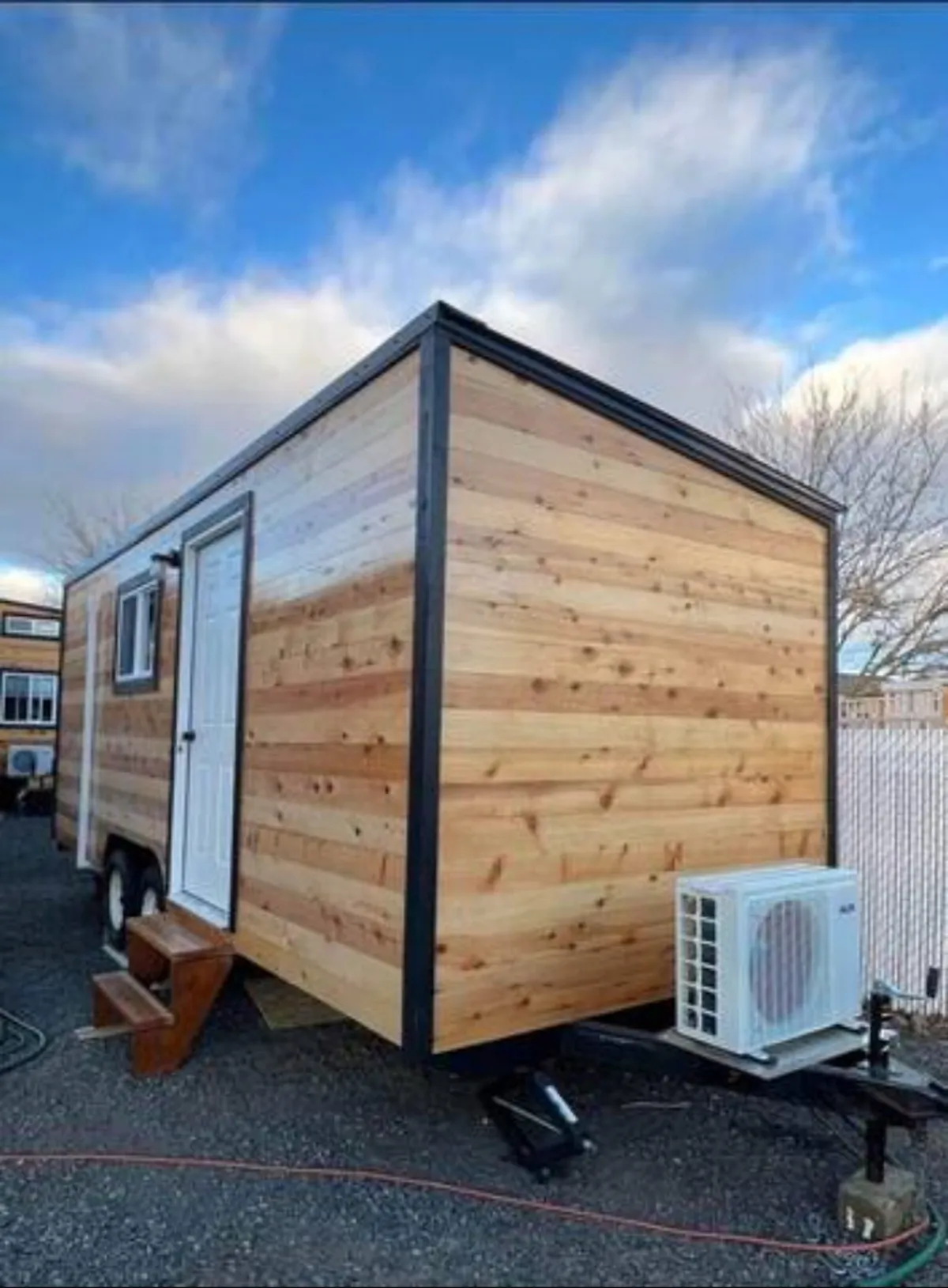 This image shows the wooden exterior of the property with a white door and an air conditioning unit, set against a backdrop of natural surroundings. The front step and rugged landscape evoke a sense of adventure and easy access to the outdoors.