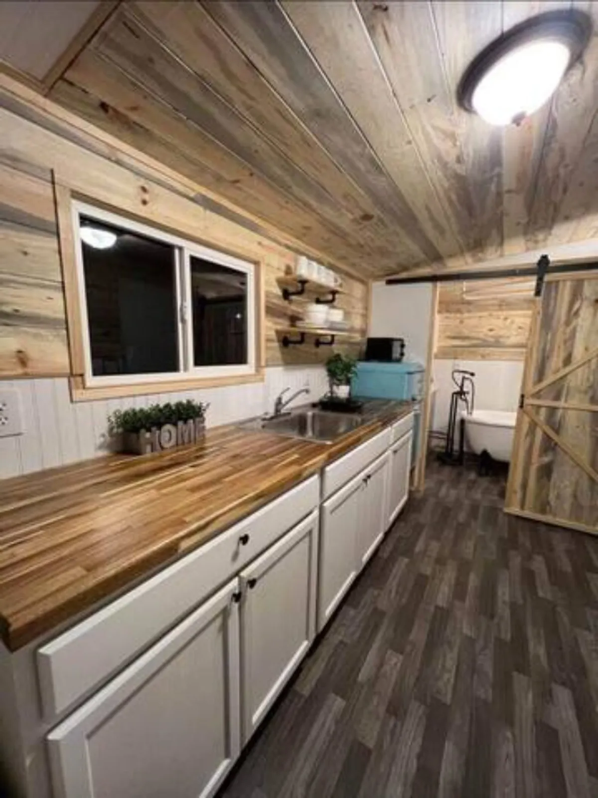 A cozy kitchen corner with a countertop, modern sink, window with natural light, a decorative plant, storage shelves, and a stylish barn door. The setting feels warm and welcoming, perfect for socializing while preparing meals.