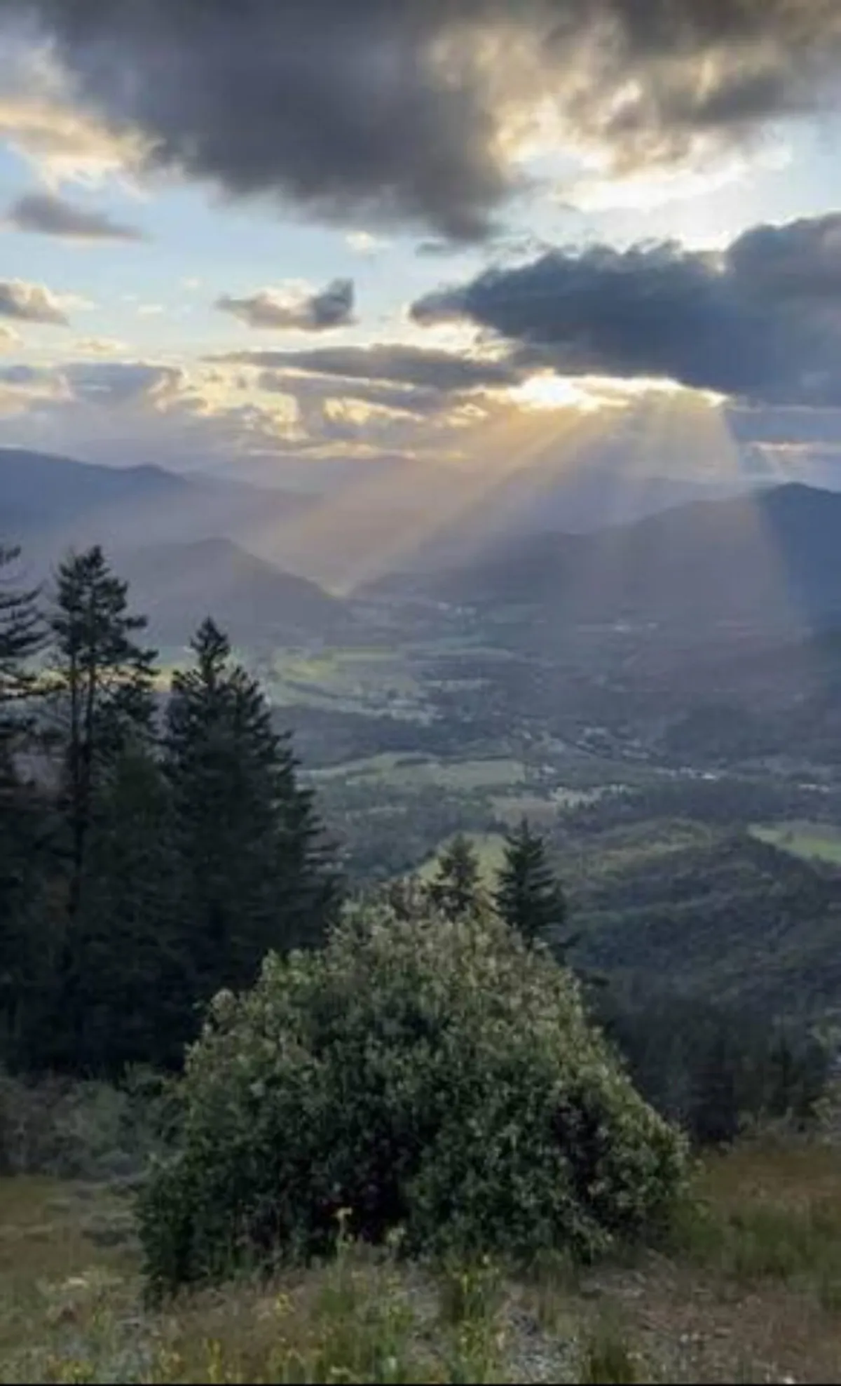 This image captures a sweeping mountain range with sun rays streaming through clouds and lush greenery in the foreground. The expansive view provides a sense of openness and the awe-inspiring natural beauty that surrounds the area, perfect for outdoor adventures.