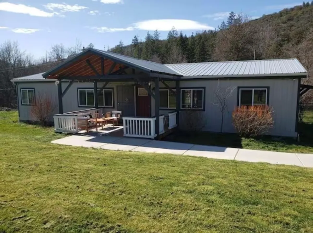 The image showcases the exterior of a charming farmhouse, featuring a spacious front porch with inviting wooden seating, surrounded by a beautifully landscaped yard. The warm sunset casts a golden glow over the scene, highlighting the peaceful and welcoming atmosphere. The setting hints at possibilities for enjoying morning coffee or relaxing outdoor meals while taking in serene valley views.