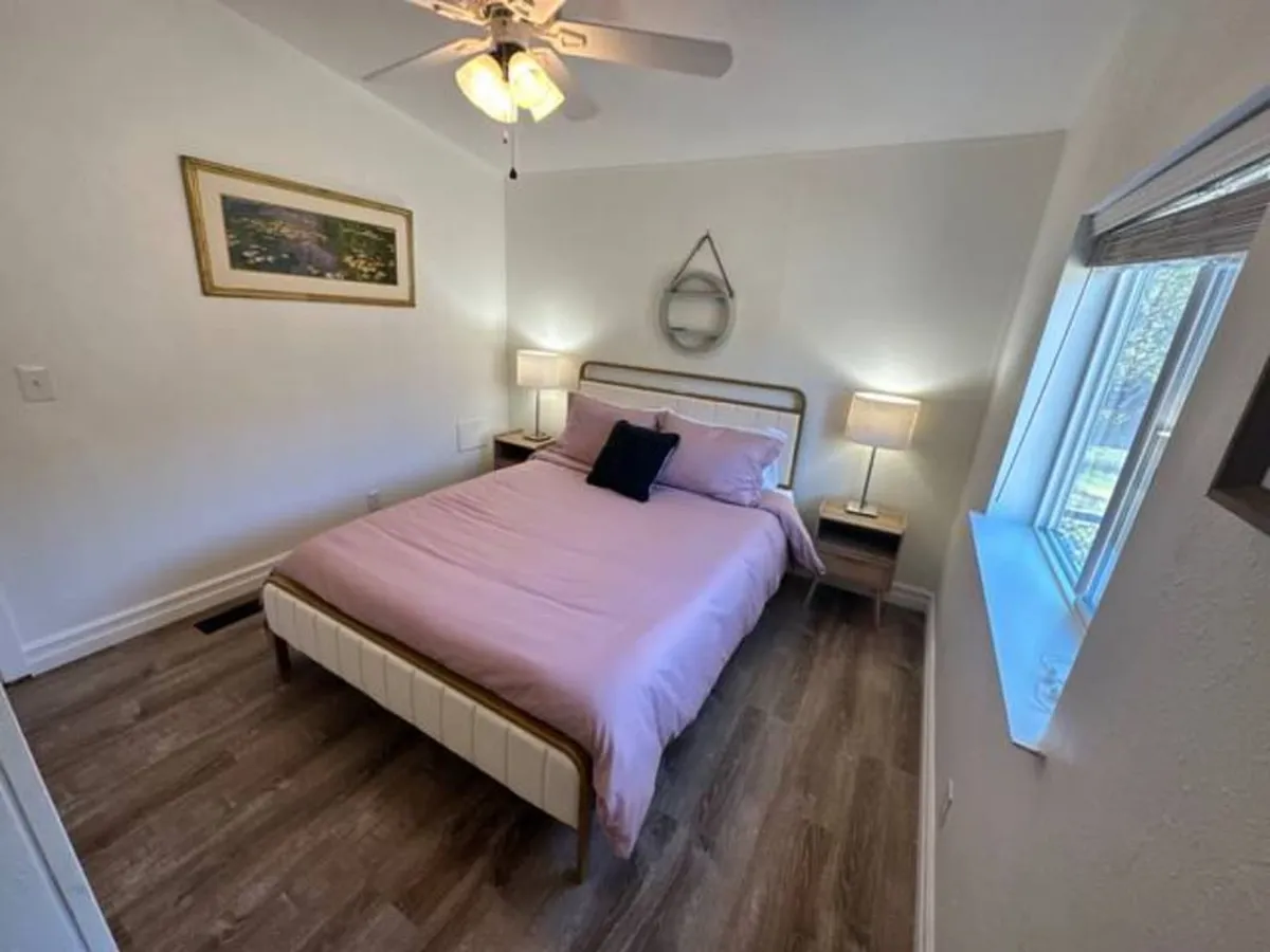This image features a cozy bedroom with a bed dressed in pink bedding, complemented by nightstands with lamps on either side, framed artwork above the bed, and a window letting in soft natural light. The mirror above the headboard adds a sense of spaciousness.