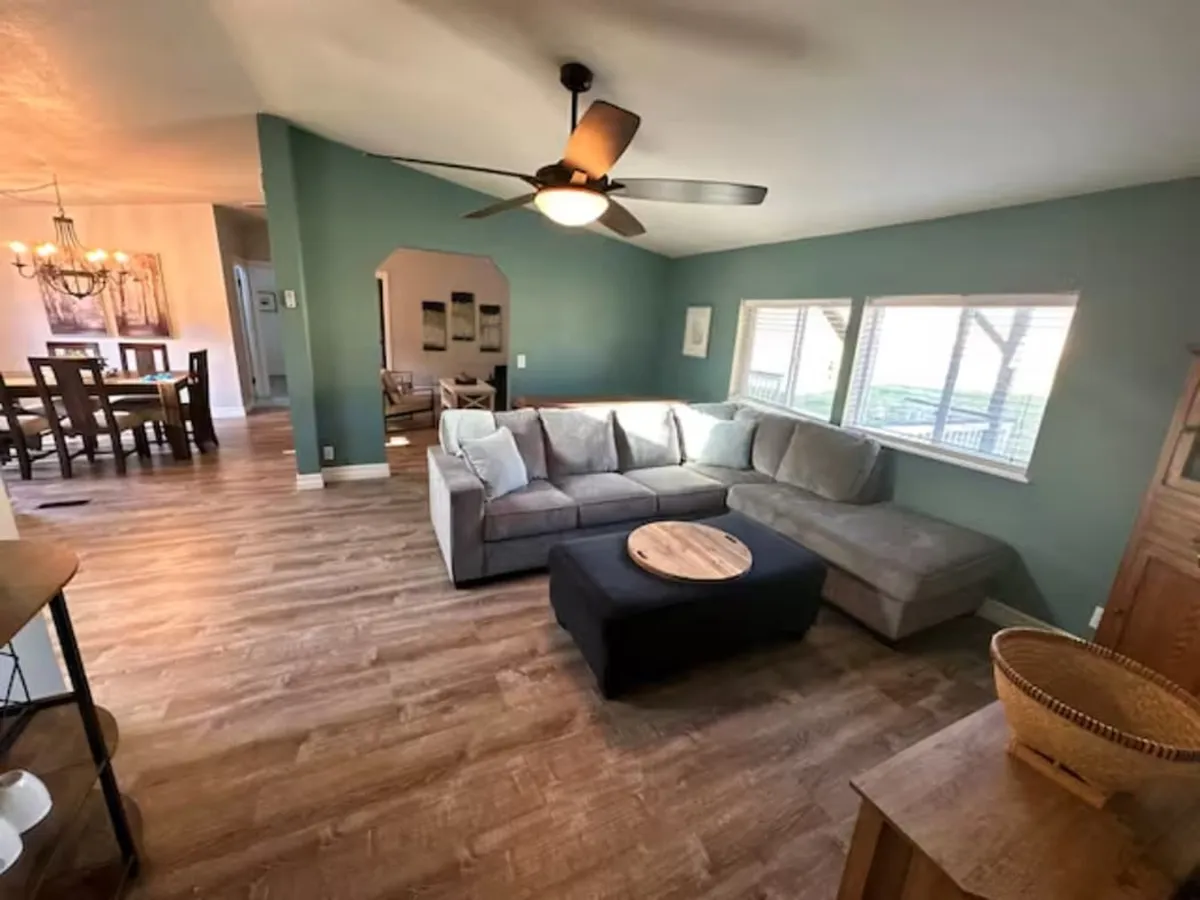 A spacious living room with a comfortable sectional sofa positioned around a coffee table, backed by large windows that let in abundant natural light and offer views of the outdoors. The room’s layout is open, with a smooth flow toward an adjacent area, creating a space that feels airy and welcoming—ideal for group gatherings.