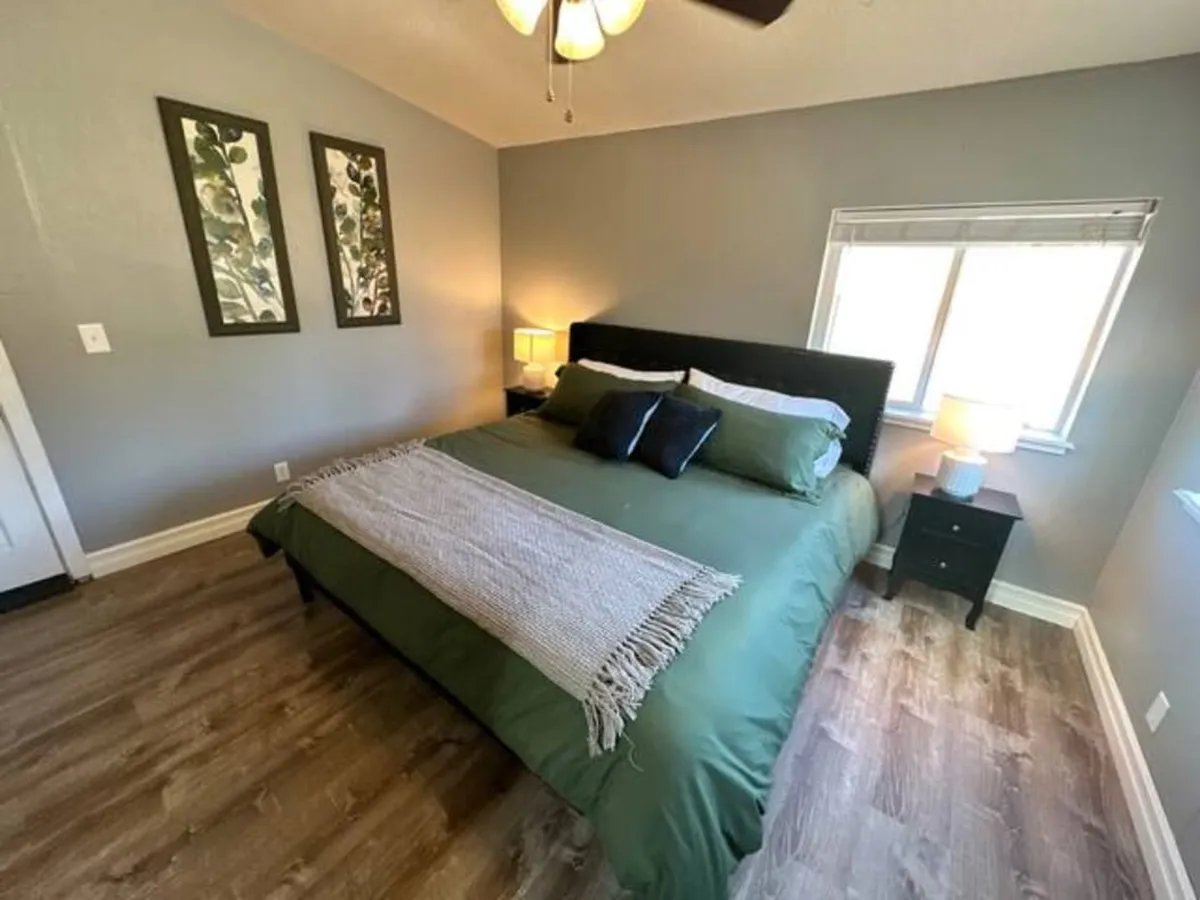 A relaxing bedroom scene showing a bed with green bedding, decorative wall art, and a lamp on the bedside table. The soft morning light filters through, creating a calm and rejuvenating atmosphere.